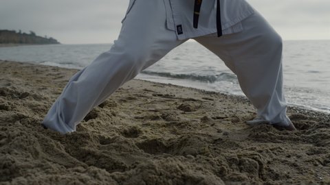 Unknown barefoot man legs making karate kicks on sand seacoast close up. Strong sportsman training feet technique martial art on morning sunlight. Unrecognizable athlete stepping on wet beach outside.