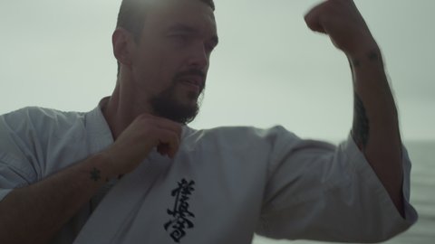 Focused athlete practicing powerful karate punches at sunset outdoor close up. Bearded middle age sportsman making fighting attack wearing kimono near sea waves. Strong man training martial arts.
