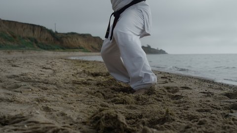 Man feet stepping on beach sand practicing karate overcast weather close up. Unrecognizable athlete doing workout on seacoast washed calm ocean waves. Barefoot sportsman exercising taekwondo outdoors.