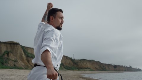 Strong fighter practicing karate on sandy beach cloudy morning close up. Bearded man training combat exercises wearing kimono overcast weather. Young sportsman making dynamic hands movement outdoors.
