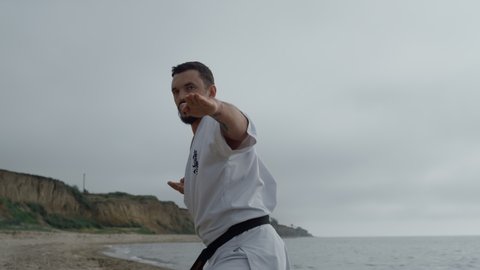 Strong sportsman practicing hands punches on gloomy beach summer morning. Focused man workout karate outside. Young athlete doing fighting exercises wearing white kimono. Martial arts concept.