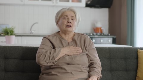 Elderly woman feels pain in her chest. Elderly woman sitting in her armchair at home is having a heart attack. Portrait of old woman with heart palpitations. Acute chest pain, heart attack.