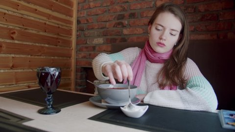 A young girl with a pink scarf sits on a sofa in a cafe and takes sour cream with a spoon and stirs it in borscht. Traditional Ukrainian food.