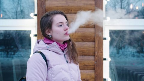 A young attractive girl in a pink jacket stands against the background of the wall of a restaurant and vapes. The concept of vaping harm to health.
