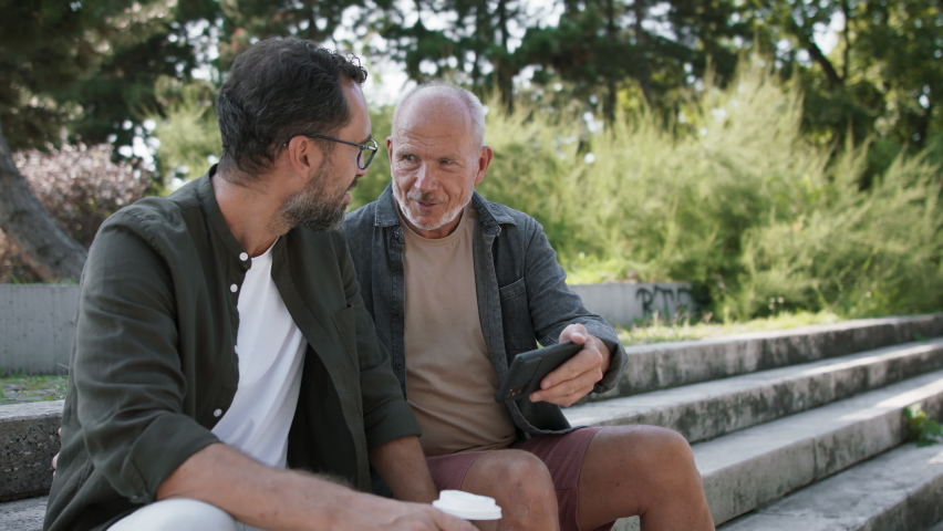 Happy senior man with his mature son sitting on stairs in park and using smartphone. Royalty-Free Stock Footage #1090175827