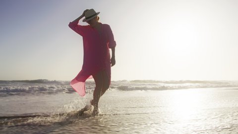 Ocean waves crashing at sandy beach and spraying in white foam and droplets. Woman in flattering pink dress and hat relaxing at golden sunset and enjoying blue blue sky view. Happiness, meditation RED