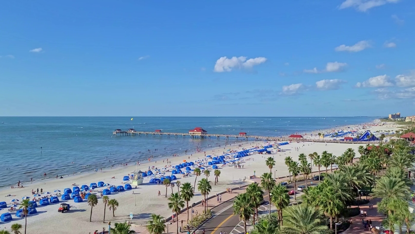 The pier of Clearwater Beach in Florida | A crowded Florida beach with people sunbathing and enjoying beach activities and seagulls flying | Also visible the palm trees and clouds in the horizon