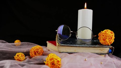 Burning candle books and thin veil with decor. Composition of burning wax candle near eyeglasses on stack of books near thin fabric with decorative wicker balls on black background