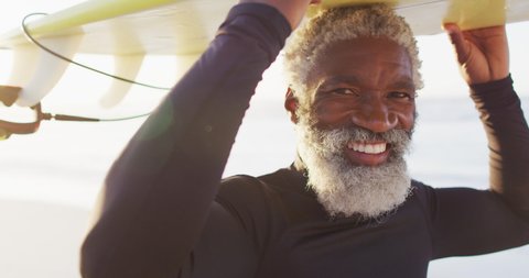 Portrait of happy senior african american man carrying surfboard on sunny beach. healthy and active time beach holiday.