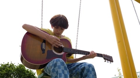 WROCLAW, POLAND - MAY 06, 2022: Teenager boy kid alone boring sit sway on swing play guitar on playground