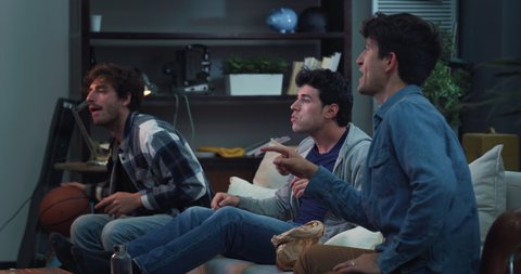Cinematic shot of young college friends football fans having fun cheering together while watching match on television sitting on sofa in living room at home.