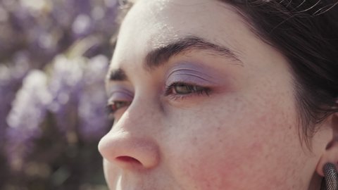 Close-up of the eyes of a beautiful woman with purple makeup. Wisteria blooms in the background.