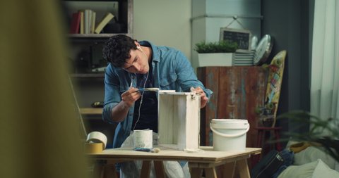 Cinematic shot of young creative man is applying white paint with paintbrush on wooden element of furniture to remodeling and renovation house by himself with diy techniques at home.