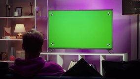 Man watching television in living room in evening. Young guy sitting on sofa against tv with chroma green screen, back view. Male person switching channels using remote controller, relaxing.