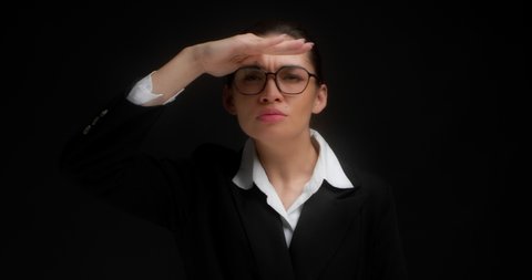 Business woman with glasses and gathered hair,covers her eyes with her hand and peering into the distance,looking far away,waiting and looking for someone on the horizon.On a black background