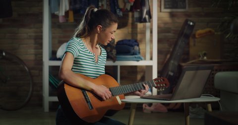 Cinematic shot of young carefree smiling woman is learning to play acoustic guitar with music teacher online streaming lesson on laptop at home.