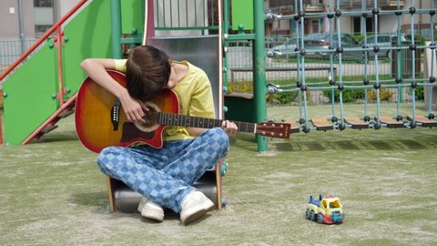 WROCLAW, POLAND - MAY 06, 2022: Boring teenager kid boy on a playground for children sit on a slide play guitar music