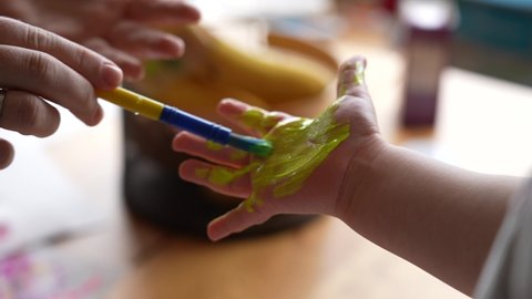 Mother or teacher preparing kid's hand to make hand prints on paper. Small child's hand and fingers covered with yellow paint. Slow motion 4k video