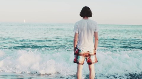Person standing in waves of the see staring at the horizon.
Long haired young caucasian man in white shirt and shorts hangs on ocean shore. Boy looks at the distance of the waters on beach. 