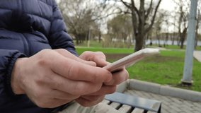 Young man is sitting on a bench in a city park, typing a text message or installing an application in his mobile phone. The smartphone is in men's hands. 4k, footage, horizontal video, close-up