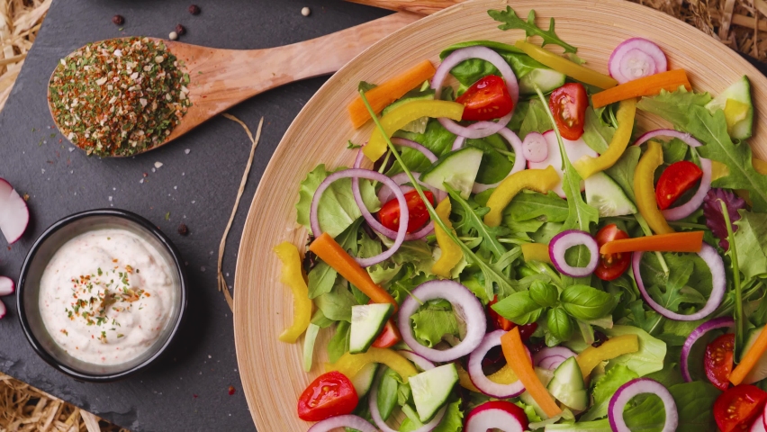 Birdseye shot of a delicious salad made with bright fresh produce, onions, tomatoes, bell peppers, served on a slate plate, spinning and a dressing on the side | Shutterstock HD Video #1090200125