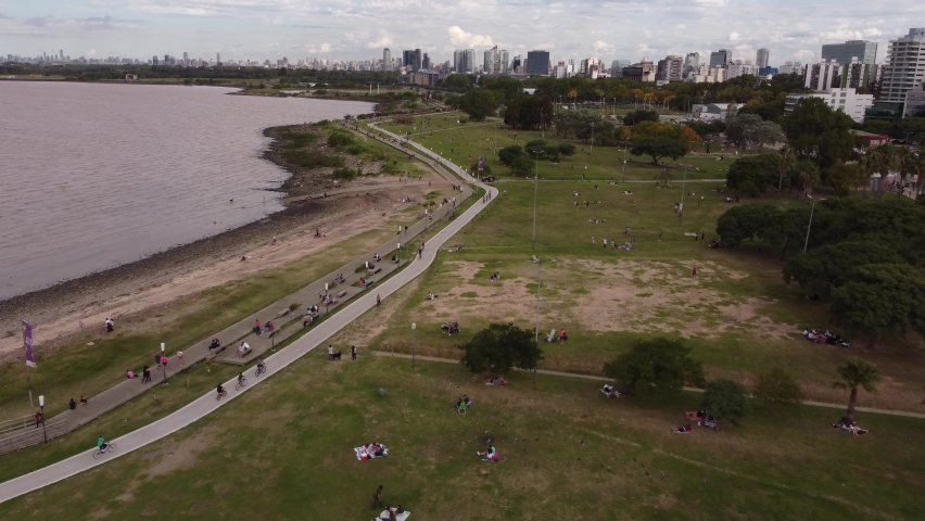 Aerial flyover Vicente Lopez Park with many people resting,walking and riding bike along river plate shore - Skyline of Buenos Aires in background