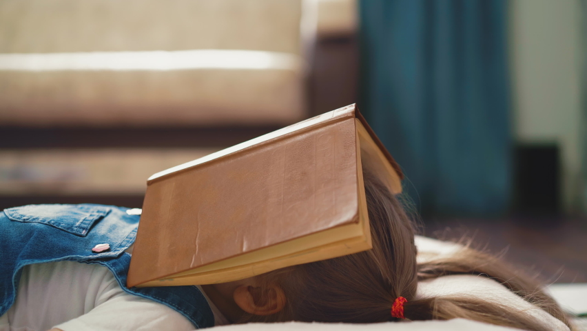 CU, Side tracking shot: little cute student girl with long hair, collected rubber bands in ponytails, lies on floor of the room, covered her face with book, tired of distance learning, closeup