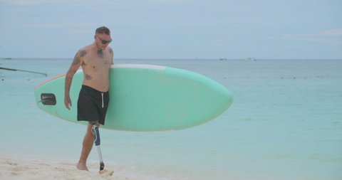 Side view of happy surfer caucasian man with disability with artificial bionic legs prosthesis holding paddle board under arm walking to ocean waters preparing for surfing on surfboard in slow motion.