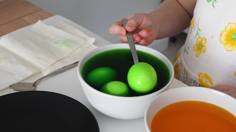 Dyed green egg in a dye bowl. A child takes a painted Easter egg from a plate with a spoon and shows it. Close-up. Family tradition of painting eggs before Easter