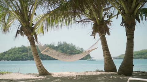 Hammock hung between two coconut palm trees on tropical relaxing beach. Perfect peaceful vacation in luxurious and beautiful island scenery. Light blue turquoise sea water. Holiday mood resting spot