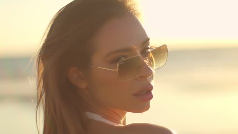 Cute pretty brunette at sunset on the ocean in sunglasses. Close-up of a beautiful woman in a white dress slow motion