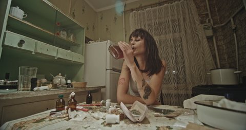 Female drug addict gulping down a bottle of booze. Female drug addict gulping down a bottle of booze as she sits at a table strewn with assorted hard drugs in a rustic kitchen