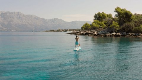 Aerial view: Man paddling on a stand up board on turquoise water. People on vacation in Croatia enjoying water sport 