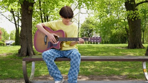 WROCLAW, POLAND - MAY 06, 2022: Lonely teenager boy sitting on a bench play guitar boring and entertaining himself