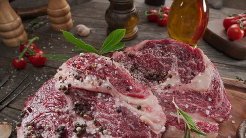 Two raw rib eye steaks with spices and herbs on a wooden kitchen table. Camera slowly moves around the kitchen table. 