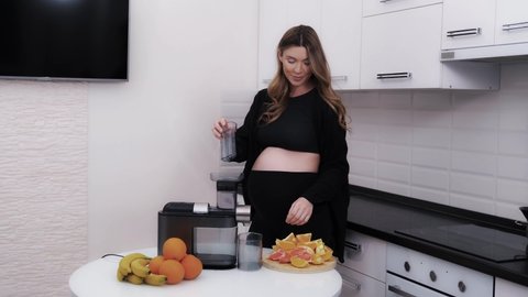 Cheerful pregnant woman using a juicer machine to make a fresh citrus drink of grapefruits and oranges. Expecting a baby requires a proper diet and vitamins. Healthy pregnancy