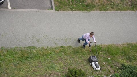 Pruszcz Gdanski, Gdansk, Poland - May 13, 2022: Woman cutting green spring grass growing on lawn in city near residential house. Aerial top view 4k video footage of person mows lawns with lawn mower
