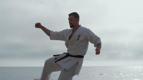 Sporty man making karate kicks on seacoast early morning close up. Serious sportsman practicing taekwondo in kimono overcast weather. Active athlete training fighting skills outdoor. Sport concept.