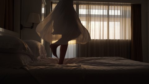 Happy woman is having fun - jumping and dancing on the bed, her skirt flying around. Low half shot, camera shows legs and skirt. Dark room, sunset sun gleam through window on background