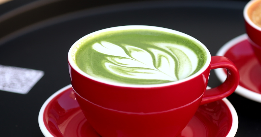 Green tea matcha latte with latte art and cappuccino in red cup on black table. Delicious milky drink on table.  | Shutterstock HD Video #1090224647