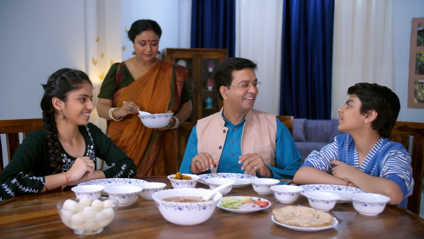 A middle-aged Bengali lady in a traditional Sari serving food to her family members - delicious food, homecooked cuisine. A modern nuclear Bengali family at dining table - family bonding, dinner time. Royalty-Free Stock Footage #1090224747