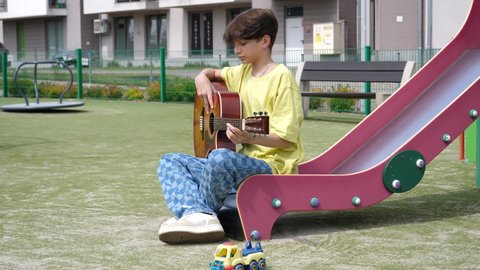 WROCLAW, POLAND - MAY 06, 2022: Boring teenager kid boy on a playground for children sit on a slide play guitar music