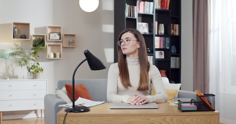 Young pretty woman in glasses sitting at desk, opening laptop computer and starting to work. Beautiful female freelancer start working day at home. Cozy room. Student studying online.