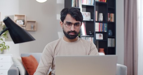 Young Arab man in glasses working on laptop computer at home during lockdown. Arabian male student texting on keyboard when studying. Freelancer work online from cozy nice room.