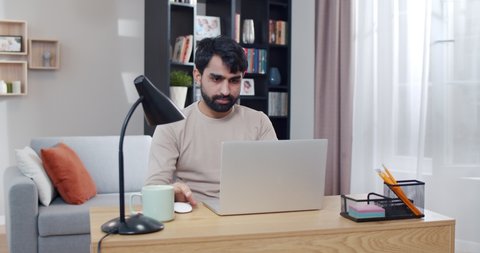 Young Arab handsome man working at laptop and getting good news or great result in job. At home. Arabian joyful male freelancer at computer. Gamer winning in game online. Lockdown concept.