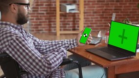 Disabled man is sitting wheelchair and talking on phone with chromakey and next to laptop with chromakey. Handicapped freelancer is having conversation on phone