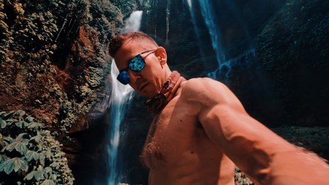 Athletic muscular man travel in tropical rainforest and enjoy beautiful waterfall against backdrop of nature background 4K Slow motion video