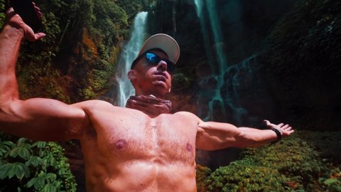 Athletic muscular man travel in tropical rainforest and enjoy beautiful waterfall against backdrop of nature background 4K