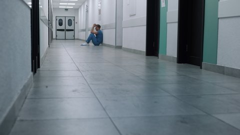 Lonely tired doctor sitting on hospital corridor floor far away. Upset overwhelmed surgeon resting alone in clinic hallway. View of unknown overworked medicine worker in empty hall after hard day.