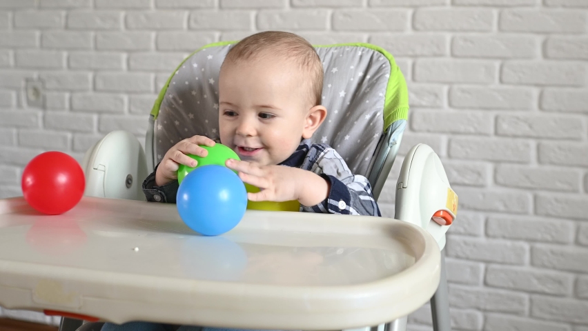 Baby boy in a plaid shirt sitting at a children's table throws multi-colored plastic balls and smiles. | Shutterstock HD Video #1090228913